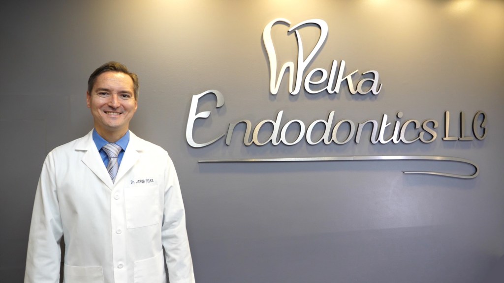 Dr. Jakub Pelka smiling in the office next to the practice logo on the wall